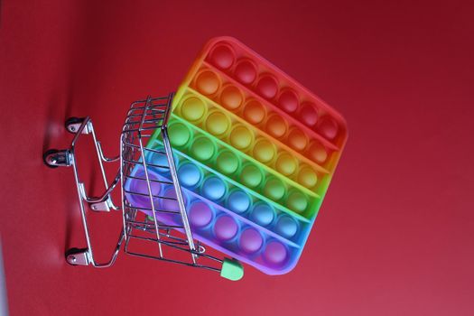 The multi-colored toy is popit simple dimple anti-stress in a basket of stroller shopping carts on a red background with a place for text copyspace. Shopping.