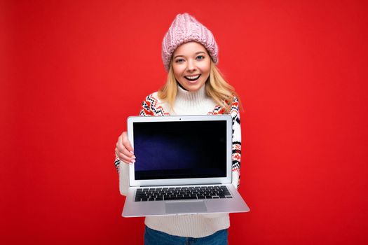 Photo portrait shot of beautiful smiling blonde young woman holding computer laptop with empty monitor screen with mock up and copy space wearing knitted winter hat and sweater looking at camera isolated over red wall background and winking.