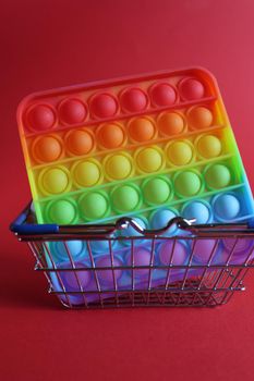 The multi-colored toy is popit simple dimple anti-stress in a basket of stroller shopping carts on a red background with a place for text copyspace. Shopping.