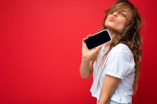 Photo of beautiful young blonde woman good looking wearing white t-shirt standing isolated on red background with copy space holding phone showing smartphone in hand with empty screen display for mockup looking at camera giving kiss.