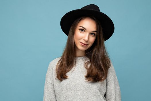 Photo portrait of young beautiful smiling hipster brunette woman in trendy pullover and black hat. Sexy carefree female person posing isolated near blue wall with empty space in studio. Positive model with natural makeup.
