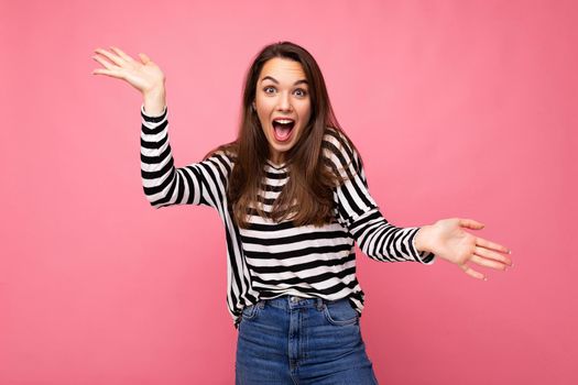 Photo of young emotional astonished shocked positive happy beautiful brunette woman with sincere emotions in casual striped pullover isolated on pink background with copy space.