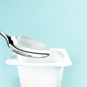 Yogurt cup and silver spoon on mint background, white plastic container with yoghurt cream, fresh dairy product for healthy diet and nutrition balance.