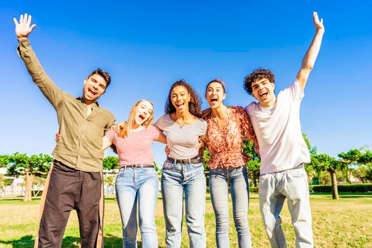 Multiracial group of young friends posing with raised open arms looking at camera embracing to each other in nature of city park. Happy diverse people having fun together enjoying life and success