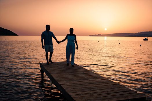 a couple seated on a wooden jetty, looking at colorful sunset on the sea, men, and women watching a sunset in Crete Greece Europe
