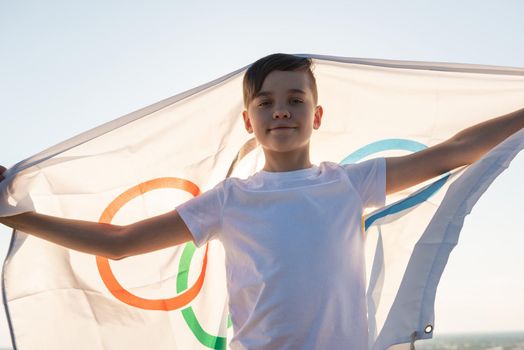 BARNAUL CITY. RUSSIA - JUNE 08, 2021: Portrait of boy waving flag the Olympic Games outdoors over cloudy sunset sky. Children sports fan. Summer olympic games concept on June 08, 2021 in Altayskiy krai, Siberia, Barnaul, Russia