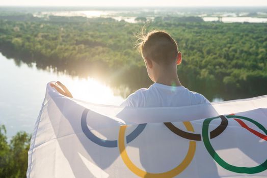 BARNAUL CITY. RUSSIA - JUNE 08, 2021: Portrait of boy waving flag the Olympic Games outdoors over cloudy sunset sky. Children sports fan. Summer olympic games concept on June 08, 2021 in Altayskiy krai, Siberia, Barnaul, Russia