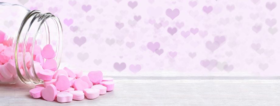 A jar of valentines heart shaped candy spilling onto a rustic wood table with heart Bokeh background.