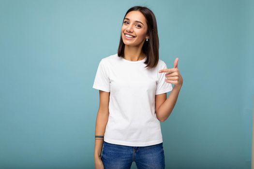Portrait of young beautiful happy smiling brunette woman wearing trendy white t-shirt with empty space for mock up. Sexy carefree female person posing isolated near blue wall in studio with free space. Positive model with natural makeup.