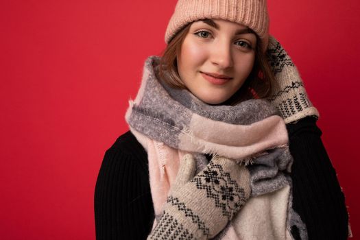 Closeup photo of beautiful happy cute young brunette woman isolated over red background wall wearing winter scarf mittens and warm hat looking at camera.