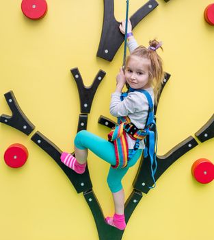 Little girl in safery harness climbs up on children's climbing wall