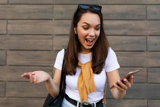 Photo of attractive shocked surprised young woman wearing casual clothes standing in the street holding and using the mobile phone looking at smartphone.