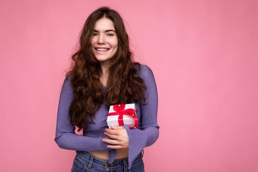 Beautiful happy young brunette woman isolated over colourful background wall wearing stylish casual clothes holding gift box and looking at camera. Copy space, mockup