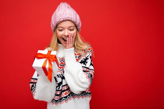 Photo shot of pretty positive surprised young blond woman isolated over colourful background wall wearing trendy outfit look holding gift box and looking at present box with red ribbon. Free space