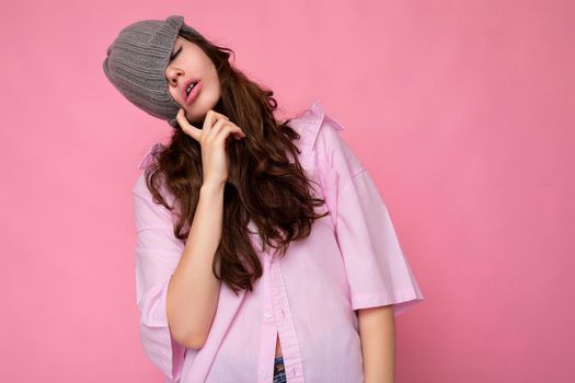 Photo of beautiful emotional young brunette woman standing isolated over pink background wall wearing pink shirt and gray hat and having fun. Copy space