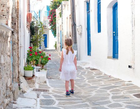 Little child girl in white dress walking the narrow alley with beautiful pink flowers between whitewashed buildings, Greece