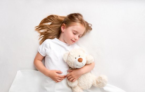 Top view of sleepy girl hugging teddy bear laying on white bed