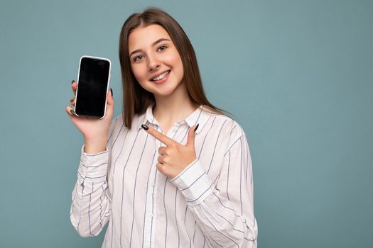 Photo of beautiful smiling young woman good looking wearing casual stylish outfit standing isolated on background with copy space holding smartphone showing phone in hand with empty screen display for mockup pointing at gadjet looking at camera.