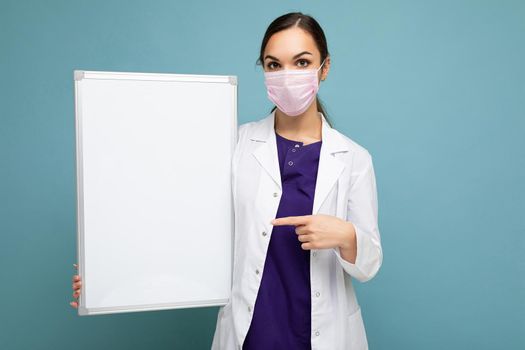 Woman doctor wearing a white medical coat and a mask holding blank board with copy space for text isolated on background. Coronavirus concept.