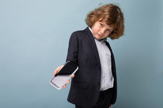Handsome positive boy with curly hair wearing suit holding phone isolated over blue background looking at camera and showing smartphone with empty display screen. Mockup