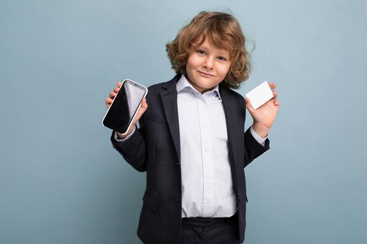 Handsome positive happy boy with curly hair wearing suit holding phone and credit card and showing mobile screen at camera isolated over blue background looking at camera. Mockup