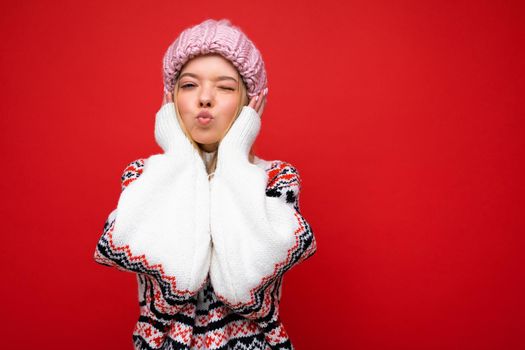 Photo of beautiful happy funny amazing young blonde woman isolated over red background wall wearing winter sweater and pink hat looking at camera and giving kiss. Empty space
