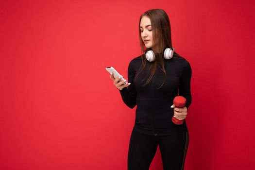 Photo of attractive positive young brunette woman wearing black sport clothes white headphones nad holding red dumbbell standing isolated over red background using mobile phone writing sms looking at gadjet display. copy space