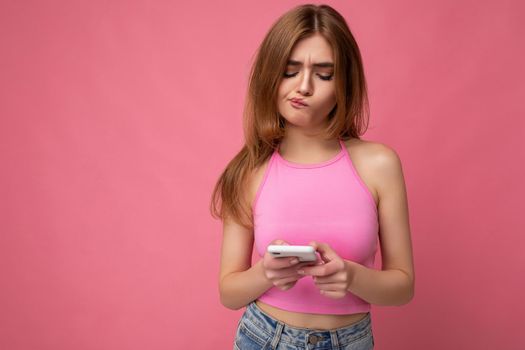 closeup Photo shot of attractive thoughtful thinking upset good looking young woman wearing casual stylish outfit poising isolated on background with empty space holding in hand and using mobile phone messaging sms looking at smartphone display screen.