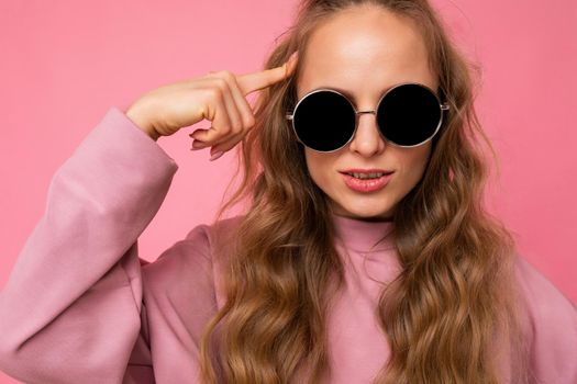 Closeup shot of beautiful serious young dark blonde curly woman isolated over pink background wall wearing casual pink sport clothes and stylish sunglasses looking at camera.