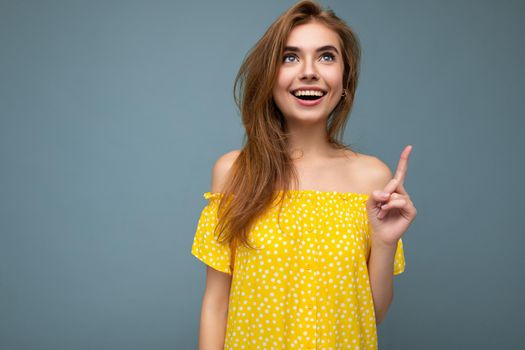 Portrait of young positive smiling sexy winsome nice attractive beautiful blonde woman wearing trendy summer yellow dress isolated over blue background with copy space and pointing up at empty space for text.
