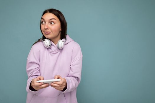 Photo of beautiful joyful smiling young woman wearing stylish casual clothes isolated over background wall holding and using mobile phone wearing white bluetooth headphones listening to music and having fun looking to the side.