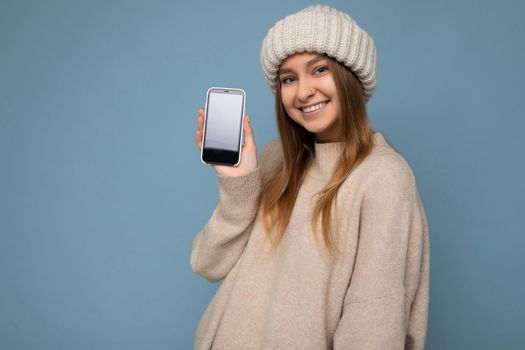 Photo of beautiful smiling positive good looking young female person wearing stylish beige sweater and beige knitted winter hat poising isolated over blue background with empty space holding in hand and showing mobile phone with empty screen for mockup looking at camera.