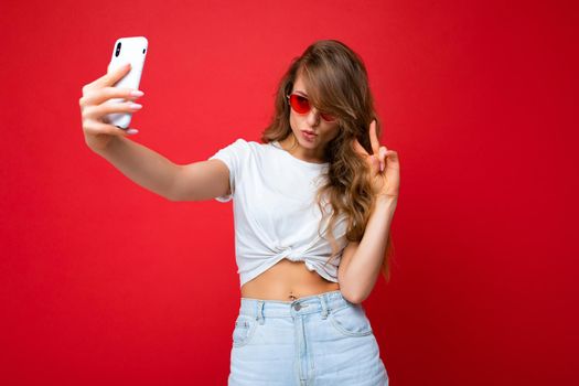 photo of amazing beautiful young blonde woman holding mobile phone taking selfie photo using smartphone camera wearing sunglasses everyday stylish outfit isolated over colorful wall background looking at device screen.