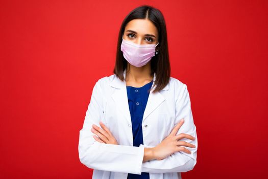 Beautiful brunette woman in virus protective mask on face against coronavirus and white medical coat isolated on the background.