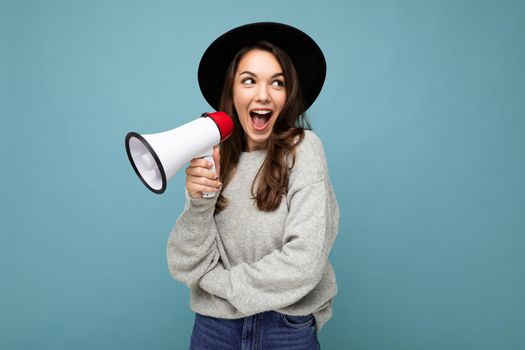 Photo of young beautiful positive happy brunette woman with sincere emotions wearing stylish black hat and grey sweater isolated over blue background with copy space and screaming in magaphone.