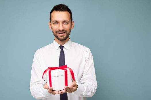 Shot of handsome positive smiling brunette unshaven young busibessman isolated over blue background wall wearing white shirt holding white gift box with red ribbon and looking at camera. Free space