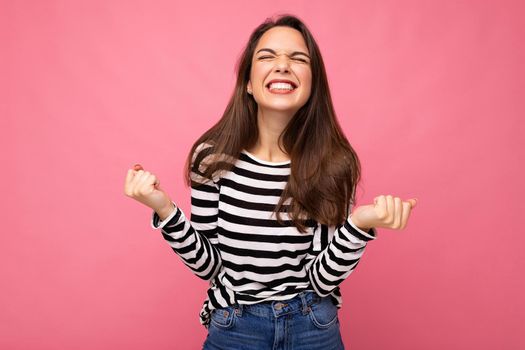 Portrait of young positive happy beautiful brunette woman with sincere emotions wearing casual striped pullover isolated on pink background with empty space and celebrating winning shouting yeah.