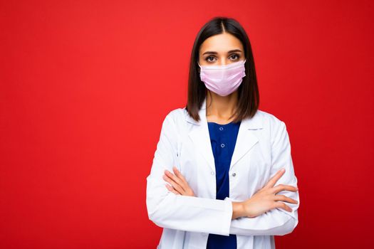 Young beautiful brunette woman in virus protective mask on face against coronavirus and white medical coat isolated on the background. Copy space