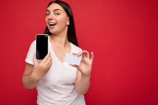 Sexy attractive happy young brunette woman wearing casual white t-shirt isolated over red background with empty space holding in hand mobile phone and showing smartphone with empty screen for mockup and credit card looking at camera.