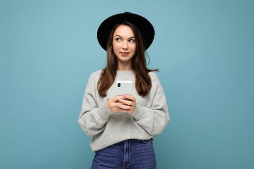 Beautiful young brunette woman thinking wearing black hat and grey sweater holding smartphone looking to the side texting isolated on background.Copy space