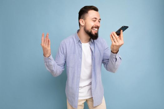 Photo shot of happy good looking young brunet bearded man wearing casual blue shirt and white t-shirt poising isolated on blue background with empty space holding in hand and communicating talking on mobile phone looking to the side.