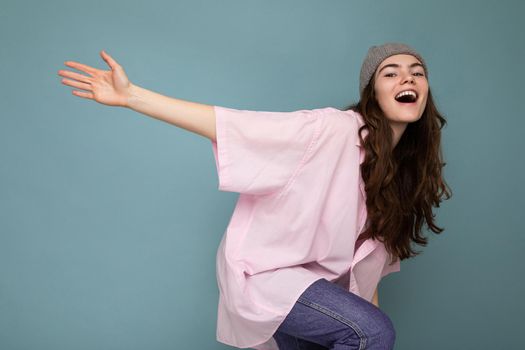 Shot of beautiful happy smiling emotional young brunette woman standing isolated over blue background wall wearing pink shirt and gray hat looking at camera and dancing.