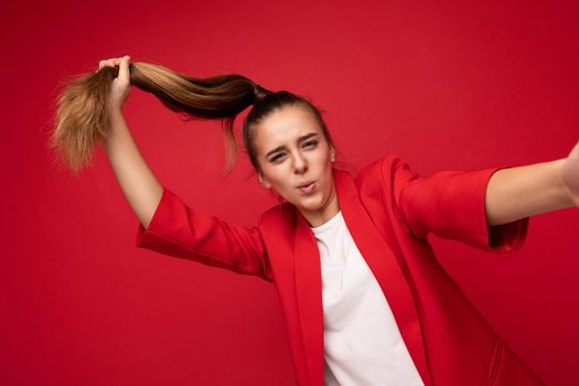 Selfie closeup photo shot of pretty positive smiling brunette little female teenager wearing trendy red jacket and white t-shirt for mockup standing isolated over red background wall looking at camera and having fun holding hair tail.