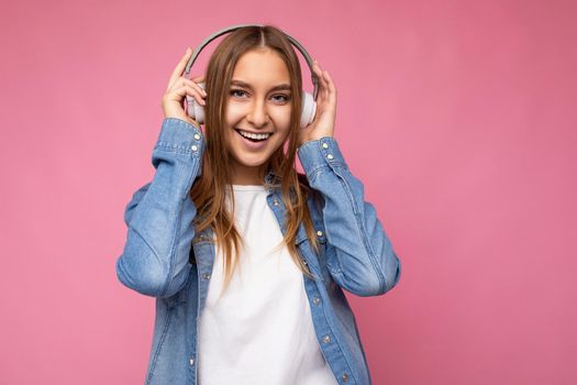 Photo of beautiful happy smiling young blonde woman wearing blue jean shirt and white t-shirt isolated over pink background wearing white wireless bluetooth headphones listening to cool music and having fun looking at camera.