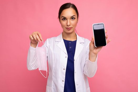 Attractive medical young woman wearing white coat holding mask showing modern mobile phone isolated over pink backgroung looking at camera.Mock up, cutout