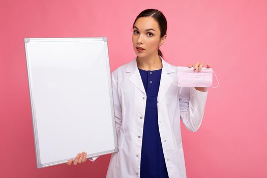 Female person doctor in a white medical coat holding blank board with copy space for text and protective mask isolated on pink background. Epidemic concept.