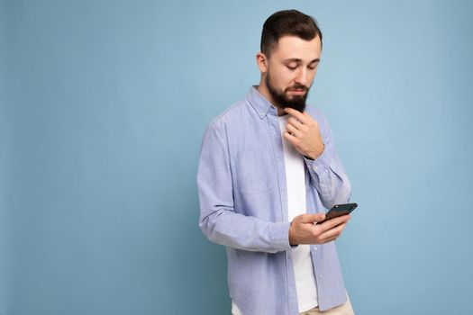 Photo shot of handsome concentrated good looking young man wearing casual stylish outfit poising isolated on background with empty space holding in hand and using mobile phone messaging sms looking at smartphone display screen.
