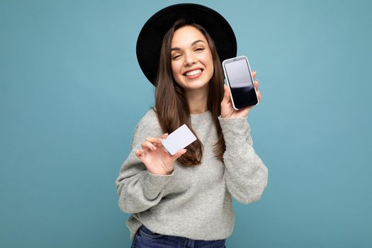 Beautiful positive smiling young brunette woman wearing black hat and grey sweater isolated over blue background holding credit card and mobile phone with empty display for mockup looking at camera.