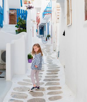 Little child girl in white dress walking the narrow alley with beautiful pink flowers between whitewashed buildings, Greece
