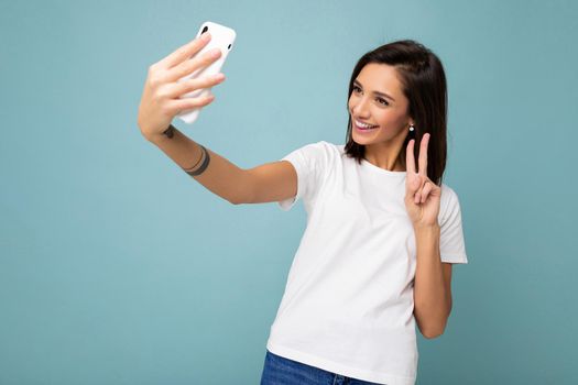 Photo of beautiful smiling happy young brunette woman wearing casual white t-shirt isolated over wall blue background holding and using mobile phone taking selfie looking at gadjet screen and showing peace gesture.